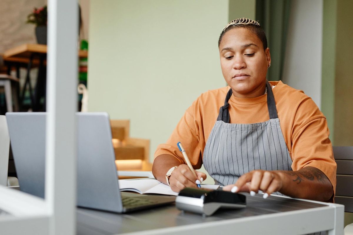 Young woman wearing apron sitting at table in front of laptop in modern cafe taking online orders fo...