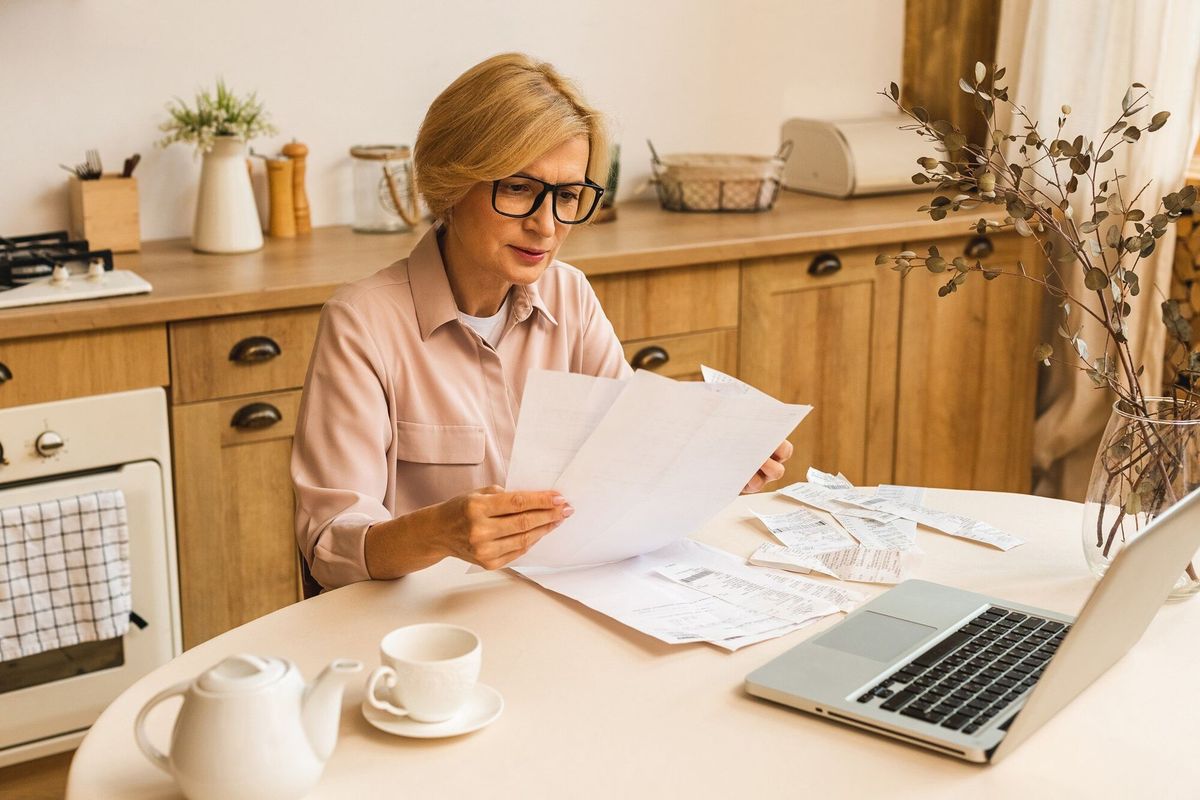 Middle aged mature senior woman holding paper bill or letter using laptop computer at home for making online payments on website, calculating financial taxes fee cost, reviewing bank account.