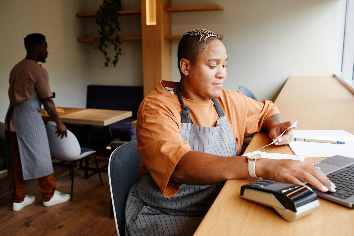Young african american woman wearing apron sitting at table in front of laptop in modern cafe taking...
