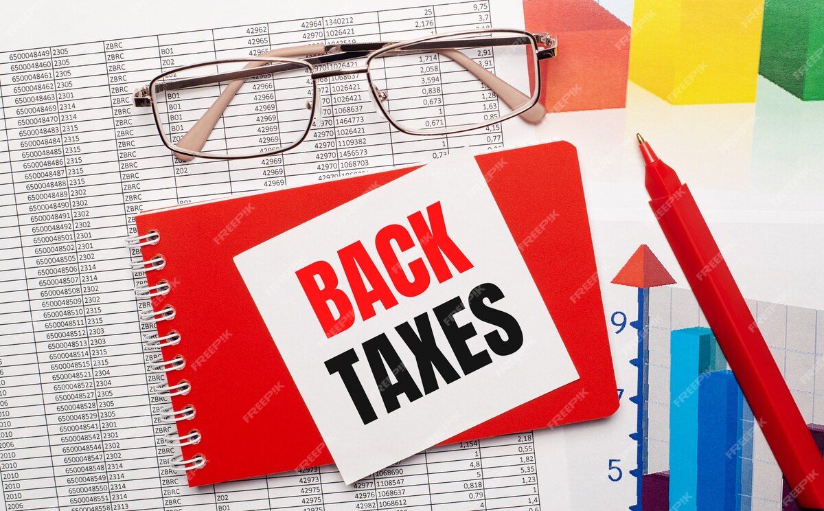 Gold-rimmed glasses, a red pen, color tables and a red notebook with a white card with the text BACK TAXES on the desktop